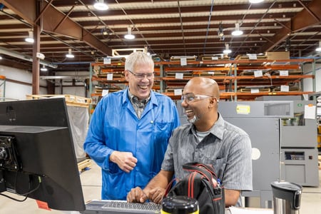 Two RIS employees smile widely as they work together on a computer. One, a white man with white hair and glasses, wears a blue ESD smock. The other, a bald South Asian man with a salt and pepper beard, wears a polo shirt and safety glasses.
