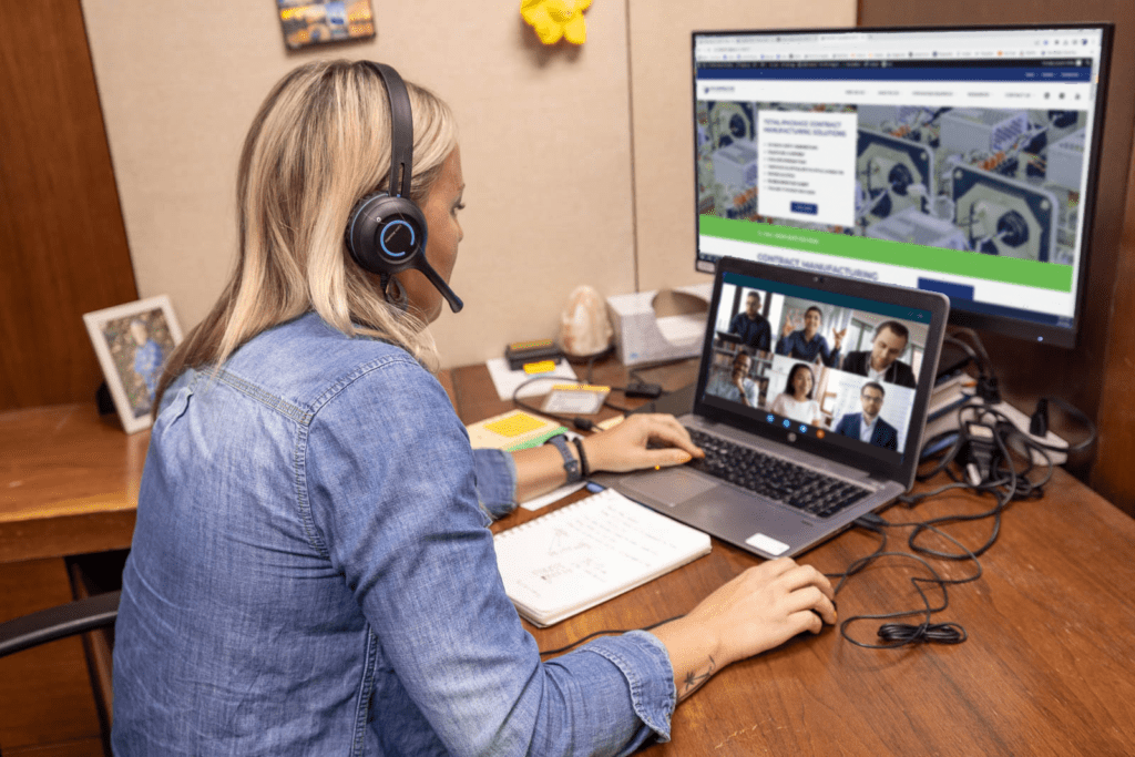 A woman with medium blonde hair, wearing a headset, faces away from the camera, sitting at a desk on a virtual meeting with several other people. At RIS, Customer Service workers work directly with teams from our OEM customers to discuss updates and changes to their accounts.